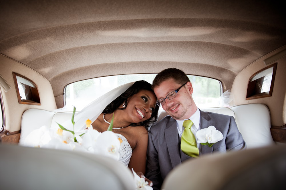 Newlywed couple sharing a tender moment in the backseat of a classic luxury wedding car, adorned with white orchids.