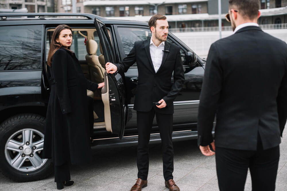 A well-dressed couple being greeted by a chauffeur next to a luxury SUV.